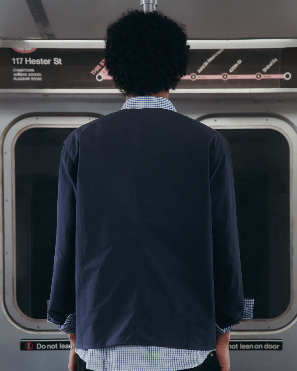 A person with curly hair stands facing a subway door, back to the camera. A subway map and station sign "117 Hester St" are visible above the door. The person wears the MAEHWA COLLARLESS SHIRT by Sundae School beneath a dark unisex mid-layer jacket.