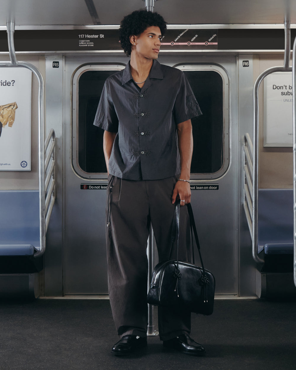 A person with short curly hair stands in a subway car, wearing wide CHARCOAL FLOWER NORIGAE LINEN TROUSERS by Sundae School, a loose dark short-sleeve shirt, and holding a black bag. Subway seats and signs are visible in the background.