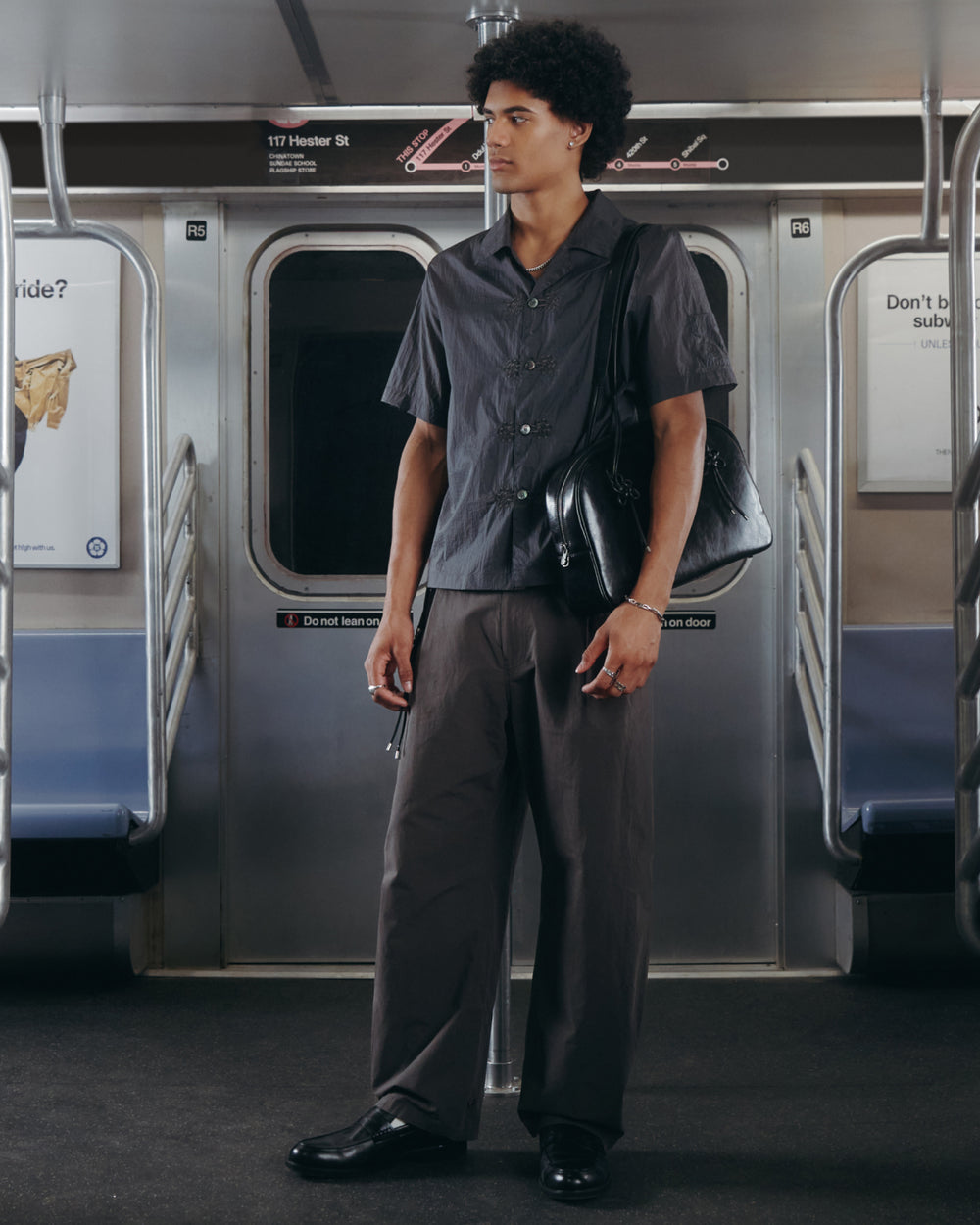 A man with curly hair stands in a subway car, dressed in dark clothes featuring Sundae School’s CHARCOAL FLOWER NORIGAE LINEN TROUSERS and holding a black bag over his shoulder. The subway seats are empty, and signs can be seen on the walls behind him.