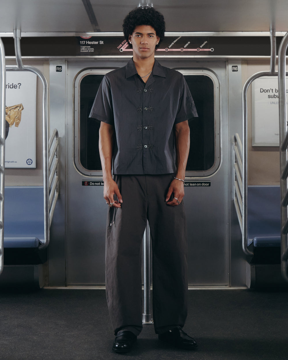 A person with short curly hair stands in the middle of a subway car, wearing Sundae School's CHARCOAL FLOWER NORIGAE LINEN TROUSERS paired with a dark short-sleeve button-up shirt. Subway seats and poles are visible behind them.