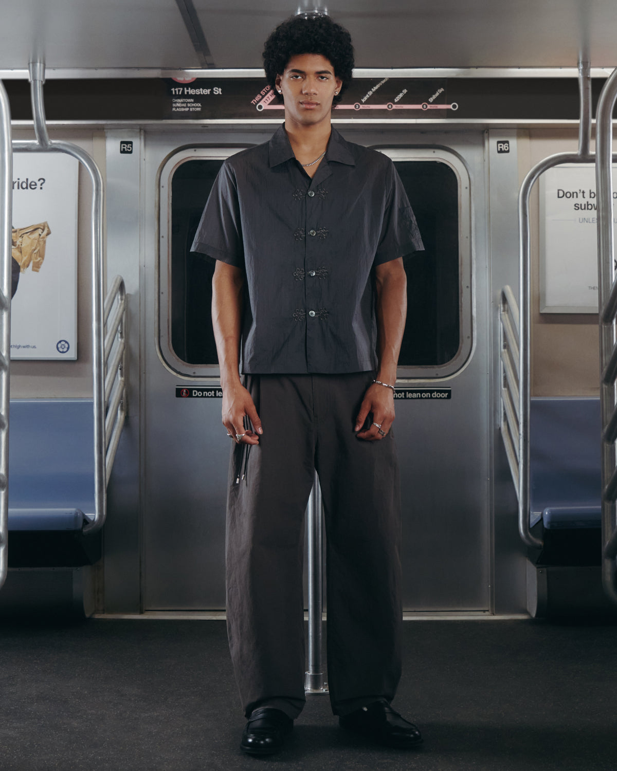 A person with short curly hair stands in the middle of a subway car, wearing Sundae School's CHARCOAL FLOWER NORIGAE LINEN TROUSERS paired with a dark short-sleeve button-up shirt. Subway seats and poles are visible behind them.