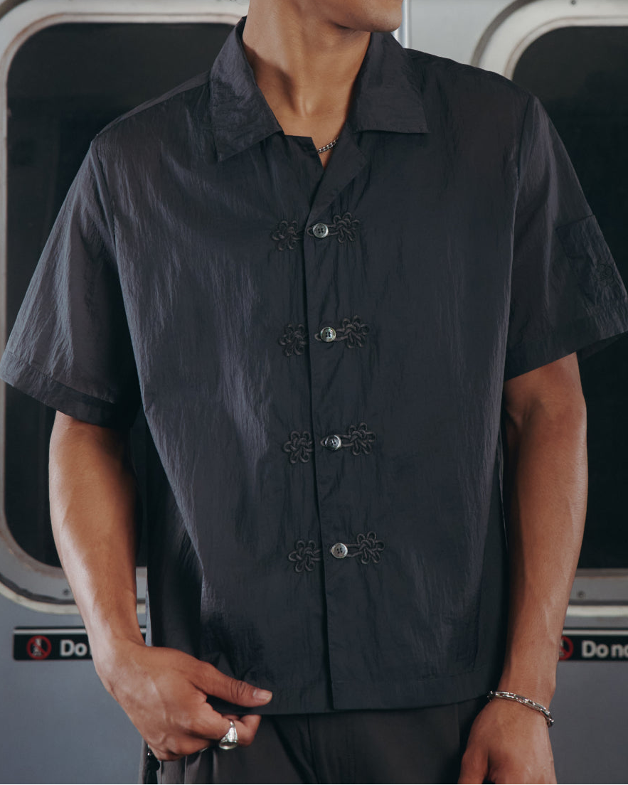 A person with curly hair, dressed in the Sundae School MAEHWA EMBROIDERED SHEER SHIRT, stands inside a subway train near the doors. A sign above reads 117 Hester St. The person looks to the side with one hand in their pocket.