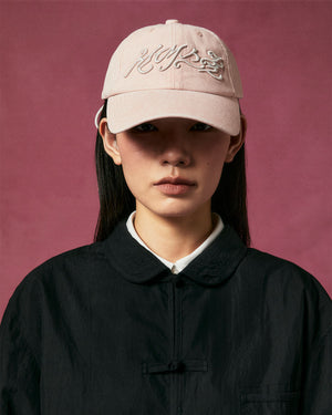 A person with straight dark hair wears a Sundae School BABY PINK FLOWER KNOT CAP, a black collared jacket, and a white shirt, standing against a dark pink background.
