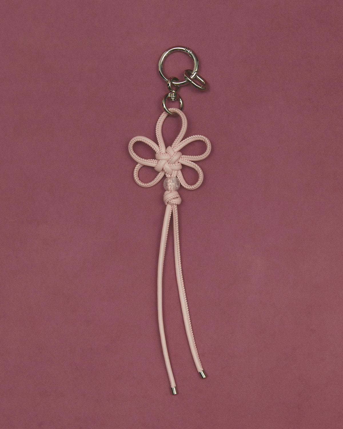 The BABY PINK FLOWER KNOT KEYCHAIN by Sundae School features a pink faux leather cord in a floral design with two dangling ends and a silver clasp, capturing Korean craftsmanship against a matching pink background.