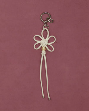 The Sundae School IVORY FLOWER KNOT KEYCHAIN, featuring a flower-shaped white faux leather cord with two long ends and a silver keyring, is shown on a mauve background.