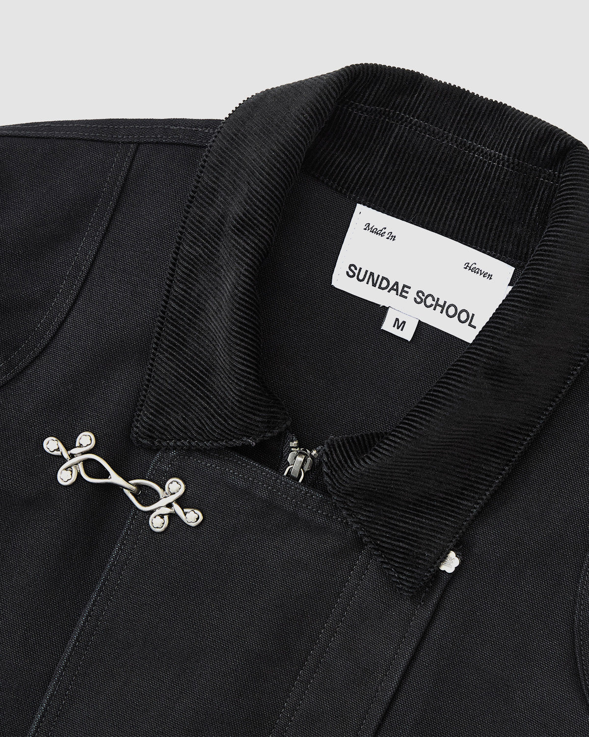 Close-up of the [PRE-ORDER] BLACK FIREMAN CLASP JACKET by Sundae School, featuring a black cotton body, corduroy collar, Mochi Floral clasp, and a label displaying Sundae School, size M, and "Made in Heaven.