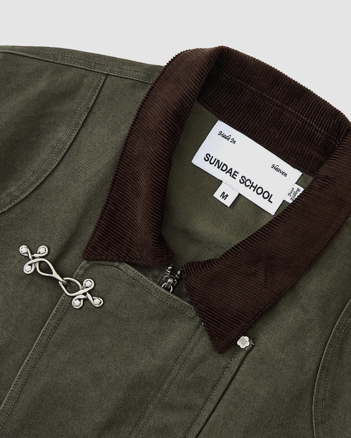 Close-up of the [PRE-ORDER] OLIVE FIREMAN CLASP JACKET by Sundae School, featuring a dark brown corduroy collar, silver branch-shaped pin, and a white SUNDAE SCHOOL label with size M and care info.