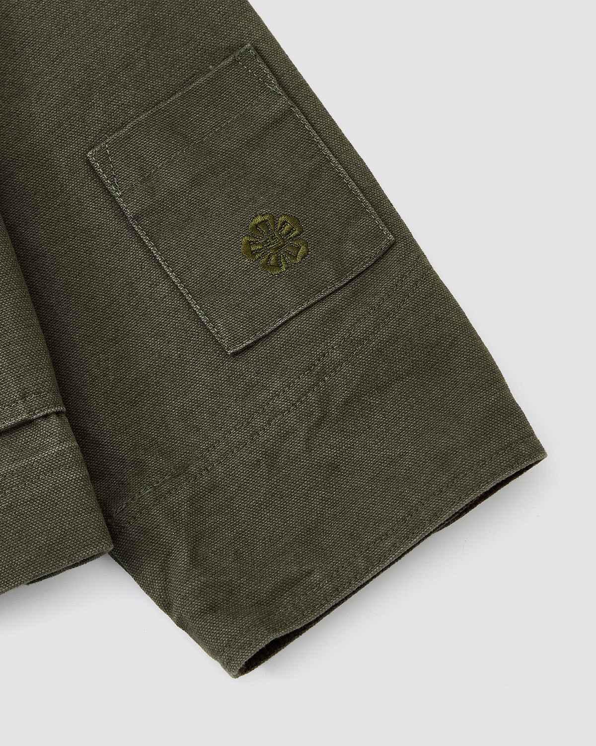 Close-up of the Sundae School [PRE-ORDER] OLIVE FIREMAN CLASP JACKET sleeve, showing a small square pocket with green four-leaf clover embroidery. The vintage cotton workwear jacket is set against a light gray background.