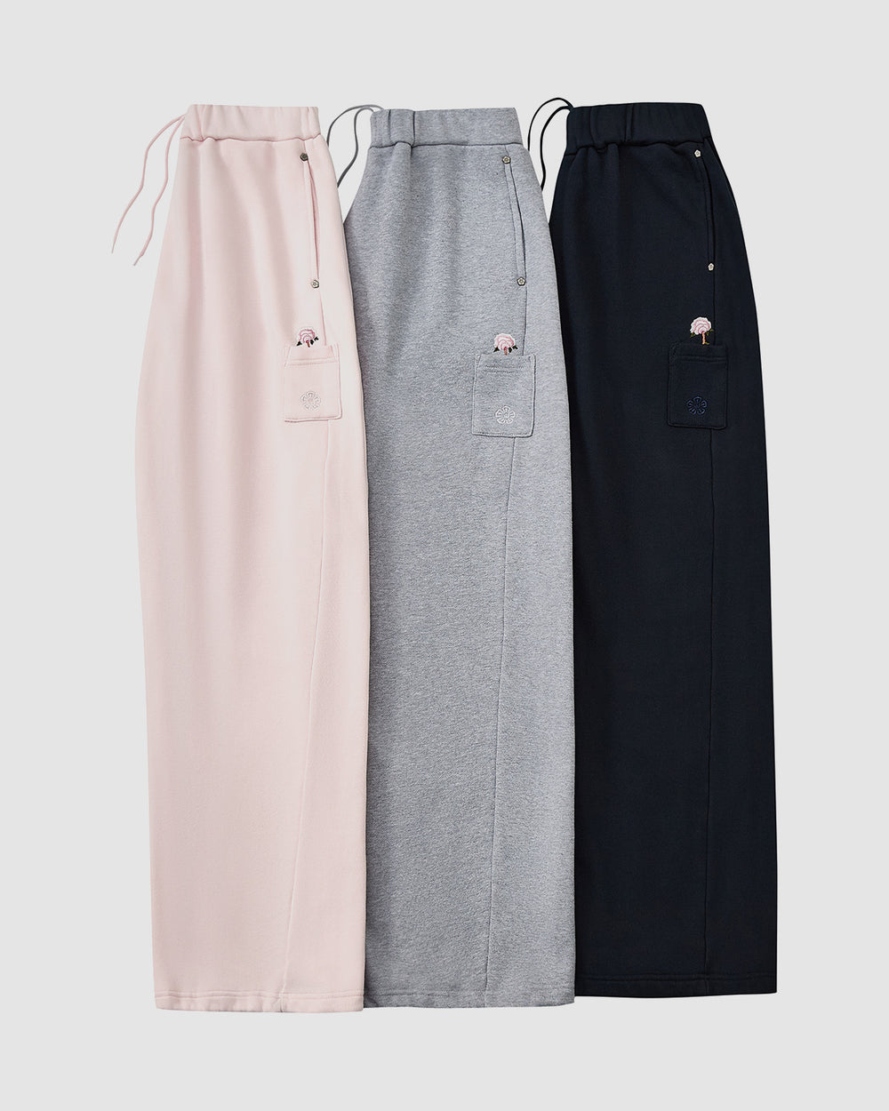 Three BABY PINK WRAP SWEATBAJI skirts by Sundae School in pastel pink, heather gray, and navy blue are displayed side by side, each with an elastic waistband, drawstring, and a patch pocket featuring subtle Korean embroidery.