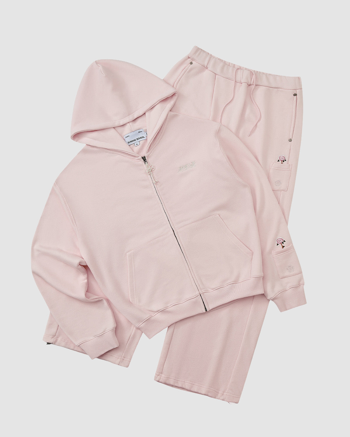 The BABY PINK WRAP SWEATBAJI set by Sundae School, featuring a light pink zip-up sweatshirt and matching wide leg sweatpants with subtle embroidery and pocket details, is laid flat on a white background for a comfortable, stylish look.