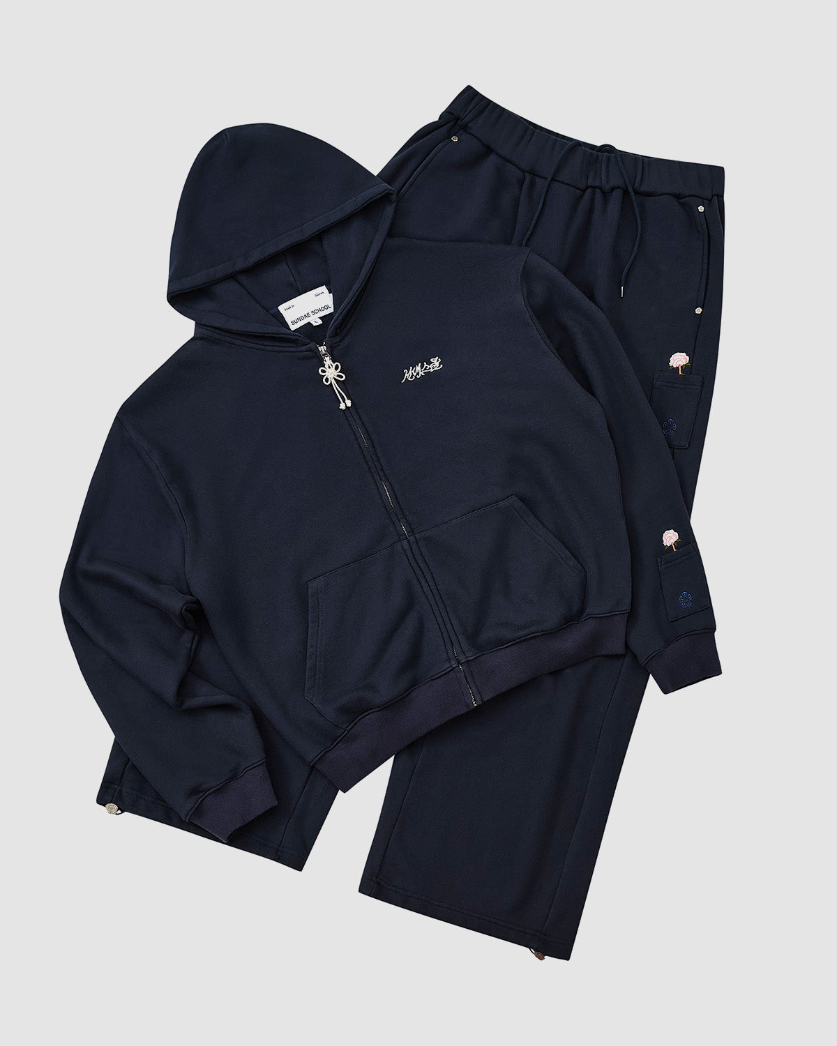 The Sundae School NAVY WRAP SWEATBAJI, a 100% cotton wide leg sweatpant with small embroidered details, is paired with a navy blue zip-up hoodie and displayed flat on a light gray background.