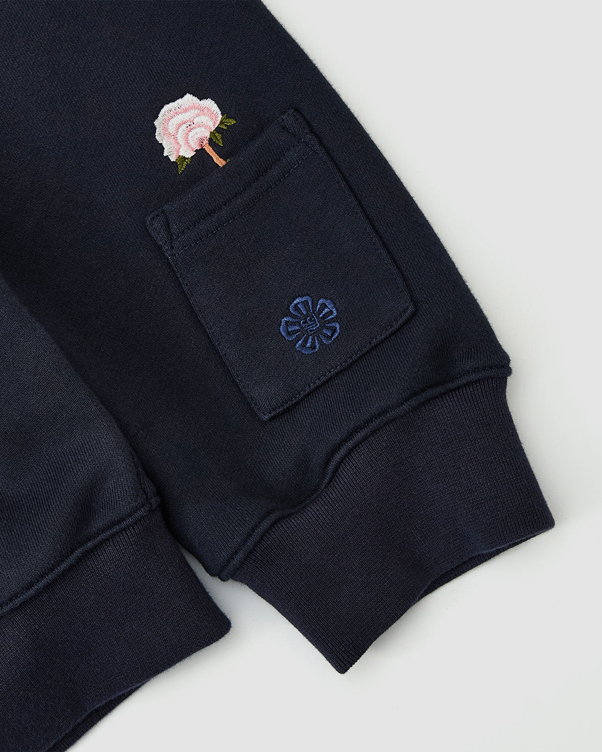 Close-up of the Sundae School NAVY FLOWER KNOT ZIP UP sleeve featuring a small pocket with mugunghwa embroidery, topped by a vibrant pink rose and green stem, capturing a modern Korean streetwear vibe.