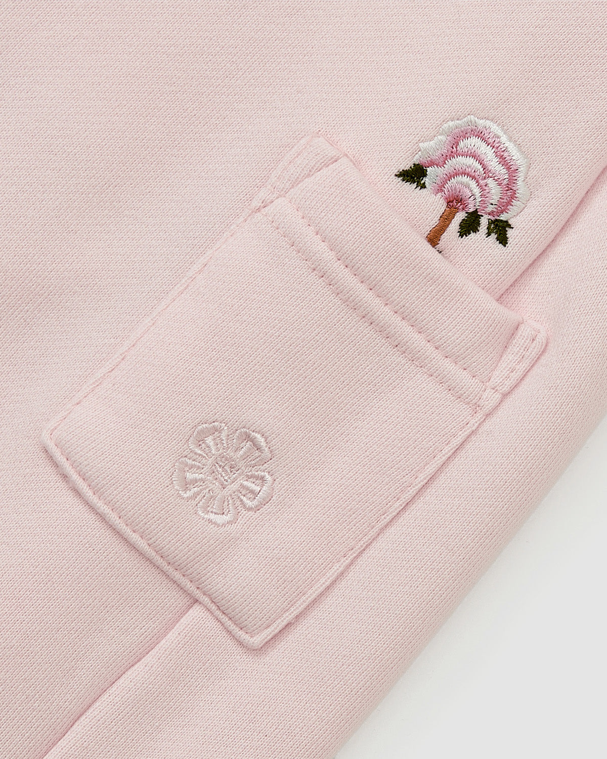 A close-up of the BABY PINK WRAP SWEATBAJI by Sundae School shows a pale pink pocket with Korean flower embroidery—an emblem on the pocket and a pink, green, and brown flower above—on these Hanbok-inspired pants.