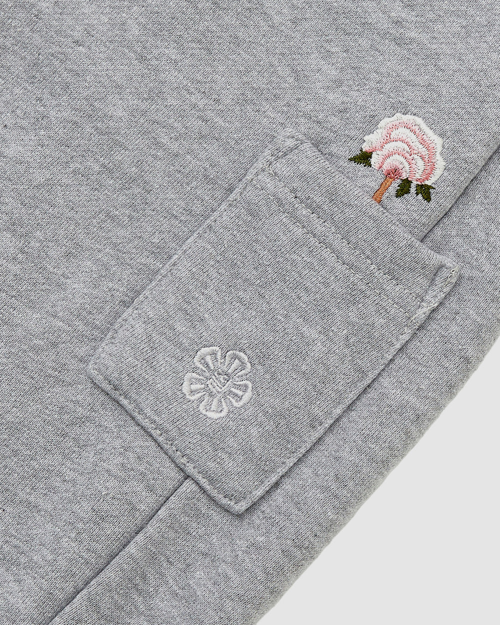 Close-up of Sundae School's HEATHER GREY WRAP SWEATBAJI: gray Hanbok-inspired pants with a small pocket adorned with white flower embroidery, topped by an embroidered pink flower with a green stem and brown outline.