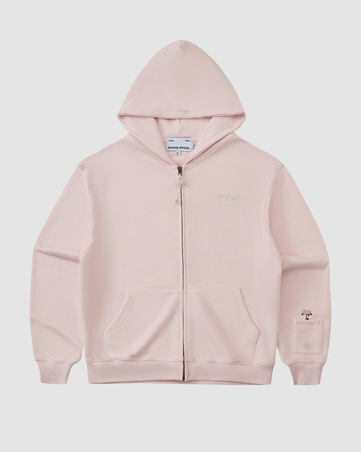 The BABY PINK FLOWER KNOT ZIP UP HOODIE by Sundae School is a light pink unisex hoodie with front pockets, subtle Korean embroidery on the chest, and a small embroidered umbrella on the left sleeve near the wrist.