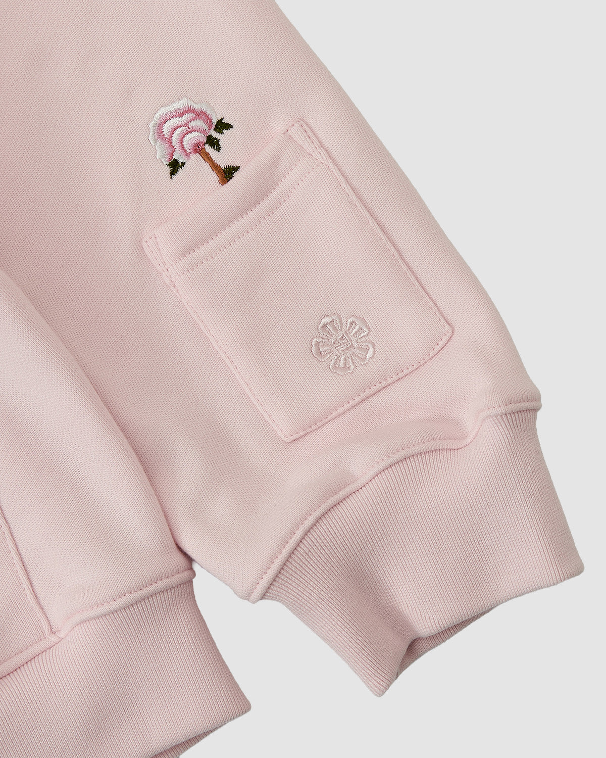 A close-up of Sundae School’s BABY PINK FLOWER KNOT ZIP UP HOODIE sleeve shows Korean embroidery of a pink Mugunghwa flower above a small pocket with a subtle embossed flower design.