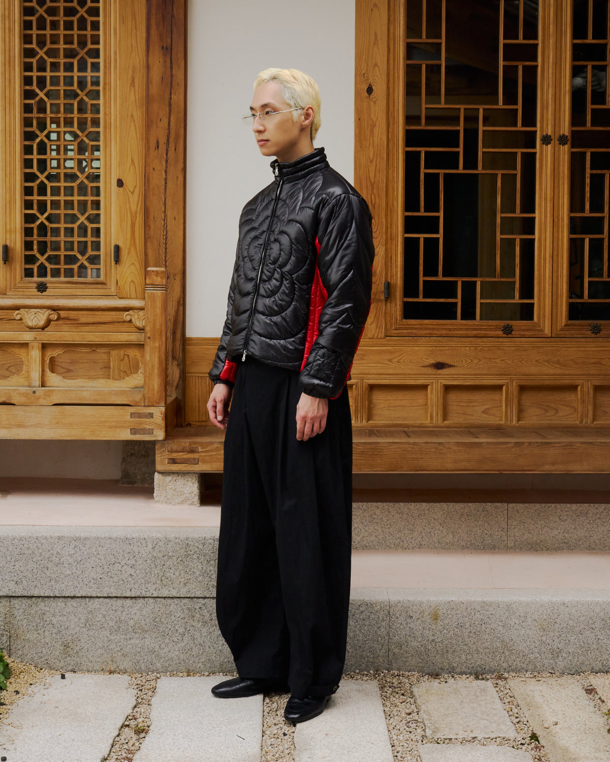 A person with short blond hair and glasses wears the Sundae School BLACK RED MOCHI SPIRAL PUFFER, loose black pants, and black shoes while standing on stone steps in front of a traditional wooden building with lattice windows.