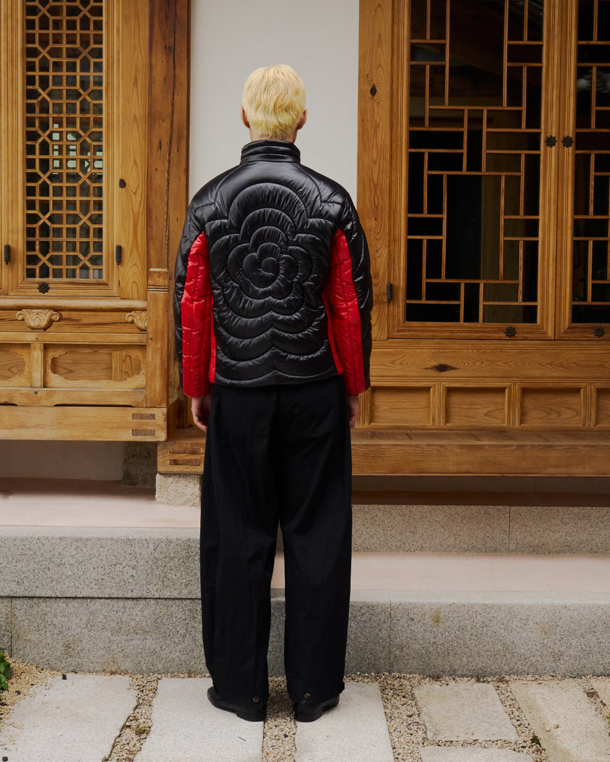 A person with short blond hair wears the Sundae School BLACK RED MOCHI SPIRAL PUFFER, paired with loose black pants and black shoes, standing in front of a wooden building with geometric window designs.