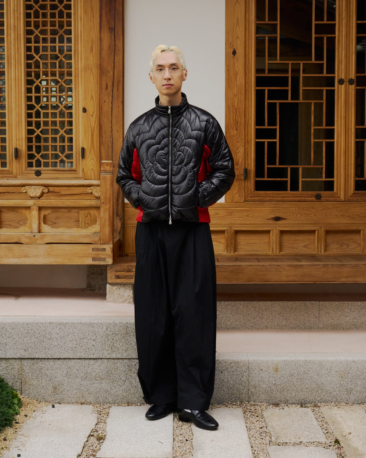 A blond-haired person with glasses stands outside a wooden building wearing Sundae School's BLACK RED MOCHI SPIRAL PUFFER, paired with wide black pants and black shoes.