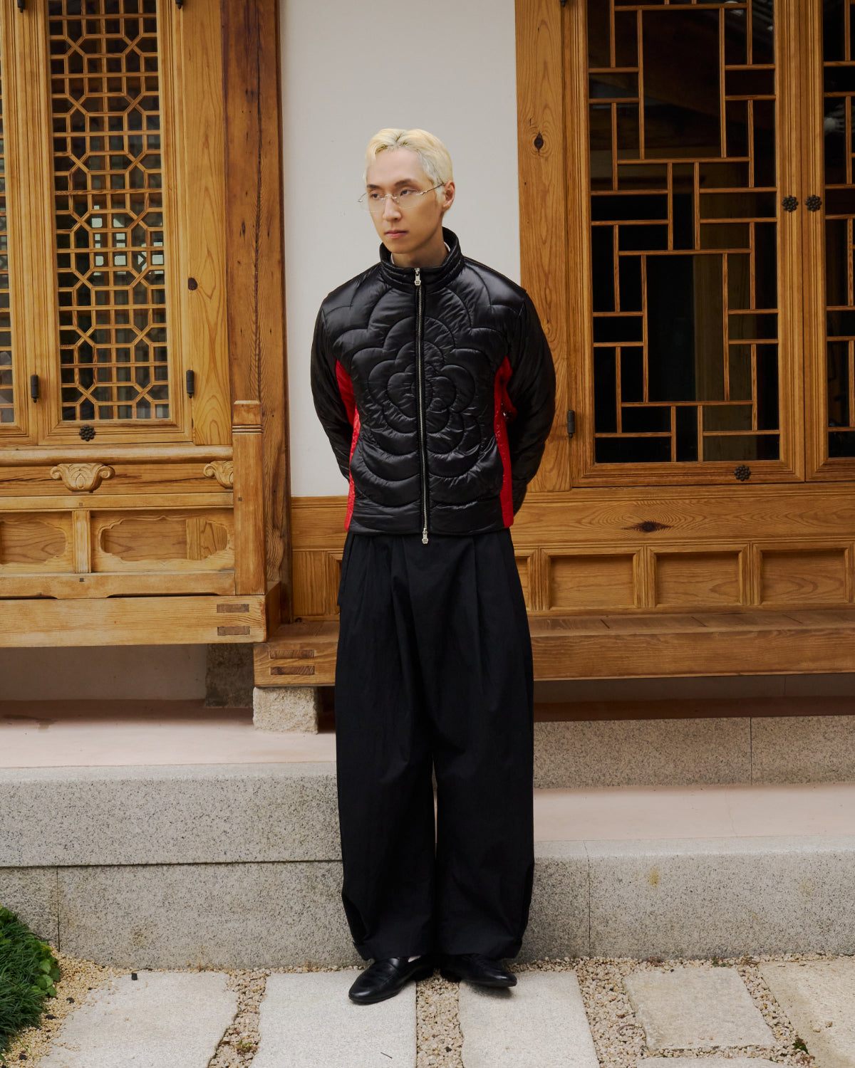 A person with blond hair, glasses, and a biker-inspired jacket stands on stone steps before ornately carved wooden doors, wearing Sundae School's BLACK RED MOCHI SPIRAL PUFFER, wide black pants, and black shoes. Hands are behind their back.