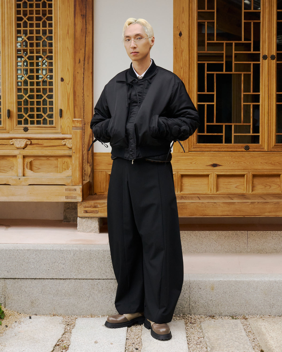 A man wearing the BLACK FLOWER CHANNELED BOMBER by Sundae School stands in front of a building.