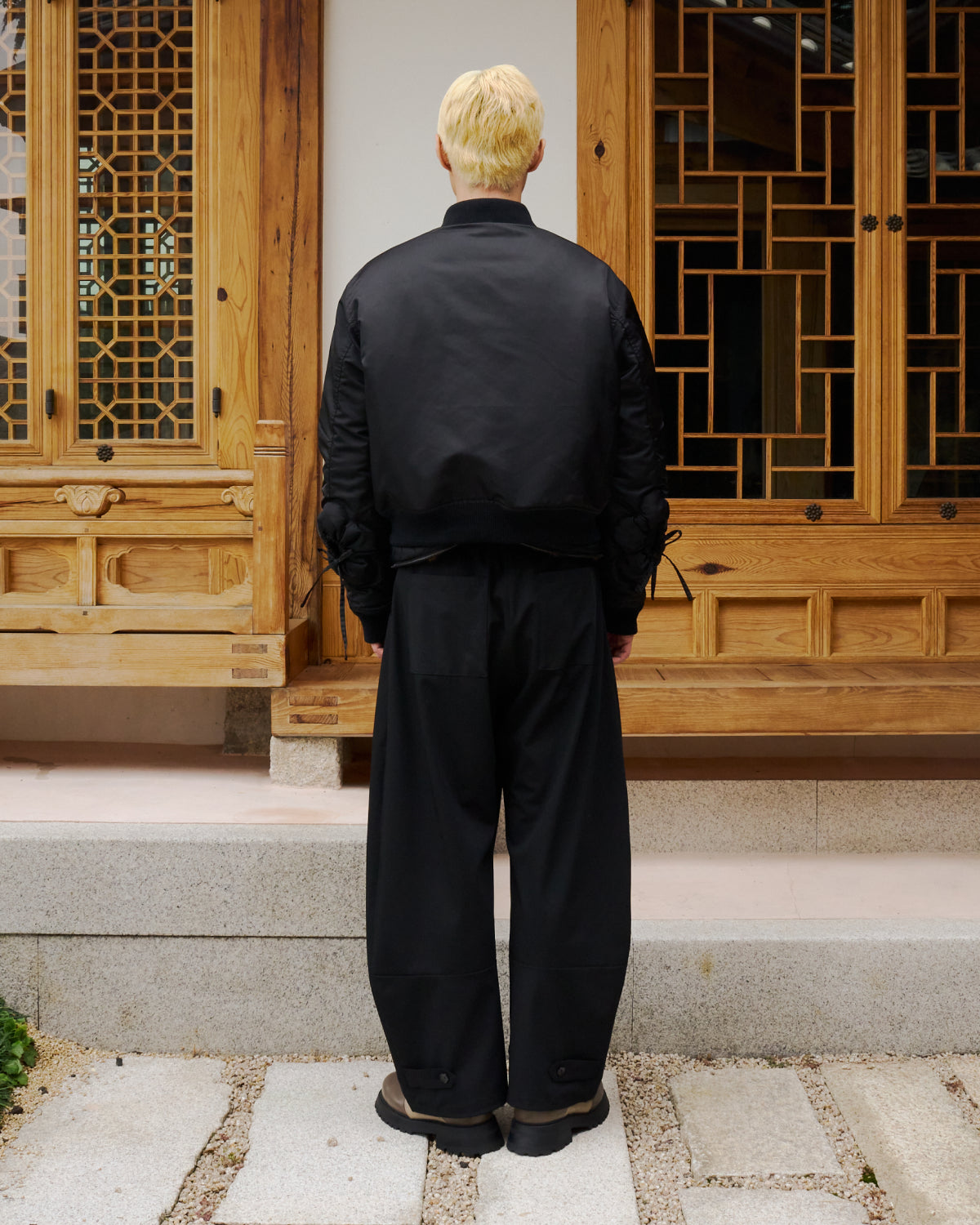 A person with short blond hair stands outdoors on stone steps, wearing the Sundae School BLACK FLOWER CHANNELED BOMBER and wide black pants, facing away in front of a traditional wooden building with intricate lattice doors.
