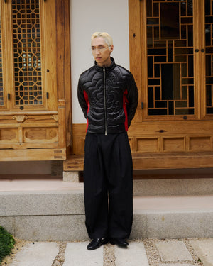 A person with blond hair, glasses, and a biker-inspired jacket stands on stone steps before ornately carved wooden doors, wearing Sundae School's BLACK RED MOCHI SPIRAL PUFFER, wide black pants, and black shoes. Hands are behind their back.