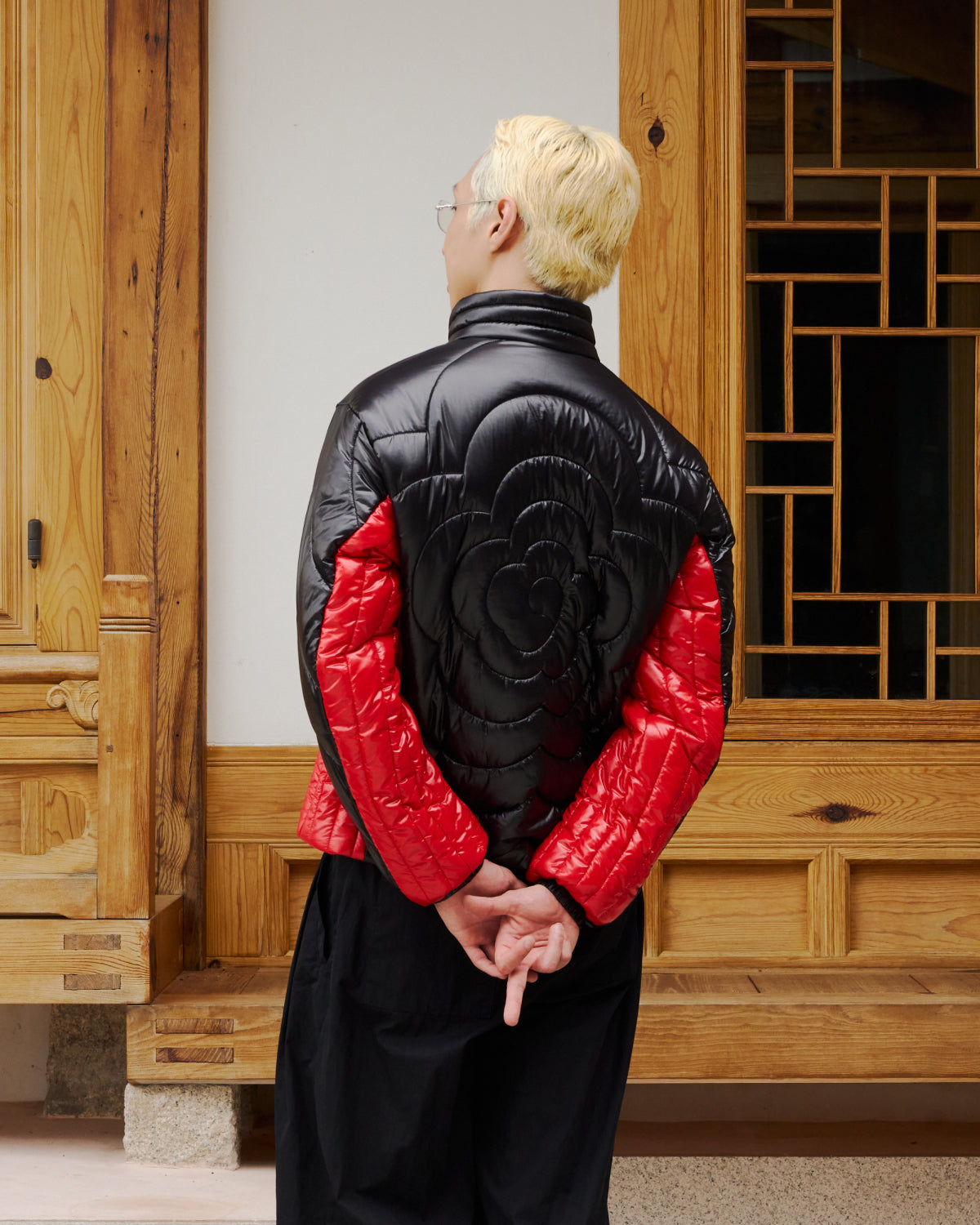 A person with bleached blond hair and glasses stands with their back to the camera, wearing the BLACK RED MOCHI SPIRAL PUFFER by Sundae School. Their hands are clasped behind their back as they stand in front of wooden doors.
