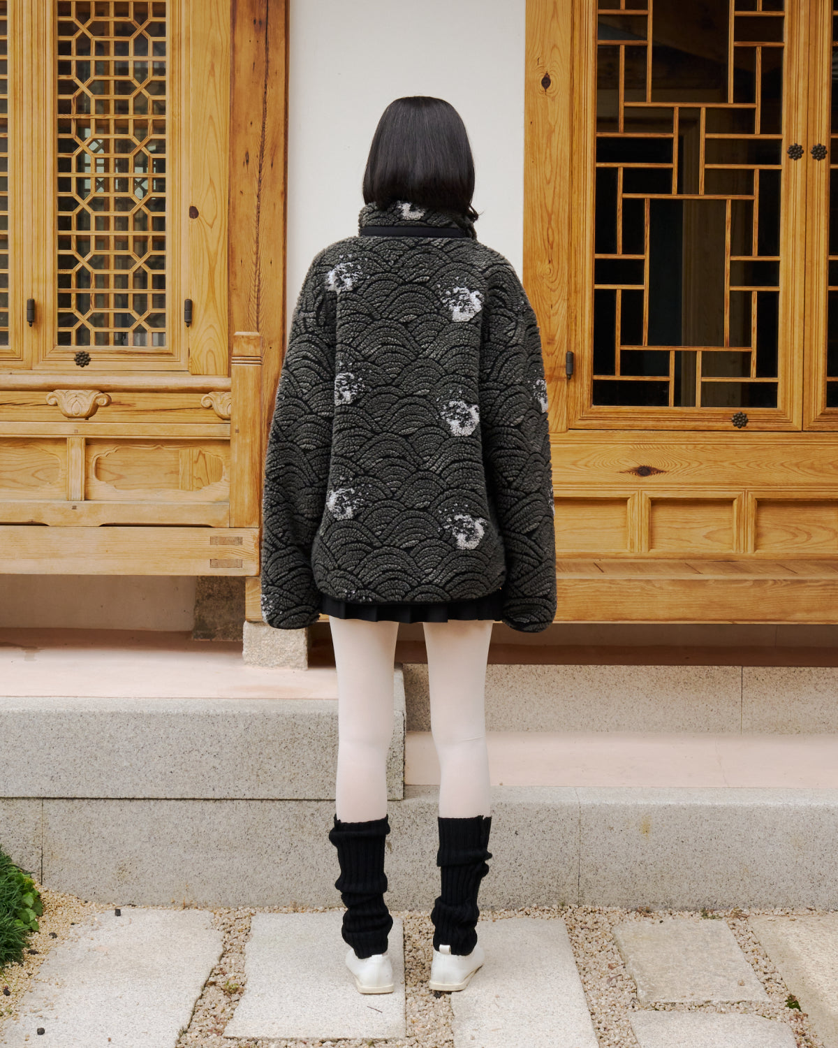 A person with shoulder-length dark hair stands facing away, wearing the Sundae School BLACK WAVE FLEECE, a short black skirt, white tights, black leg warmers, and white shoes on stone steps before a wooden building.