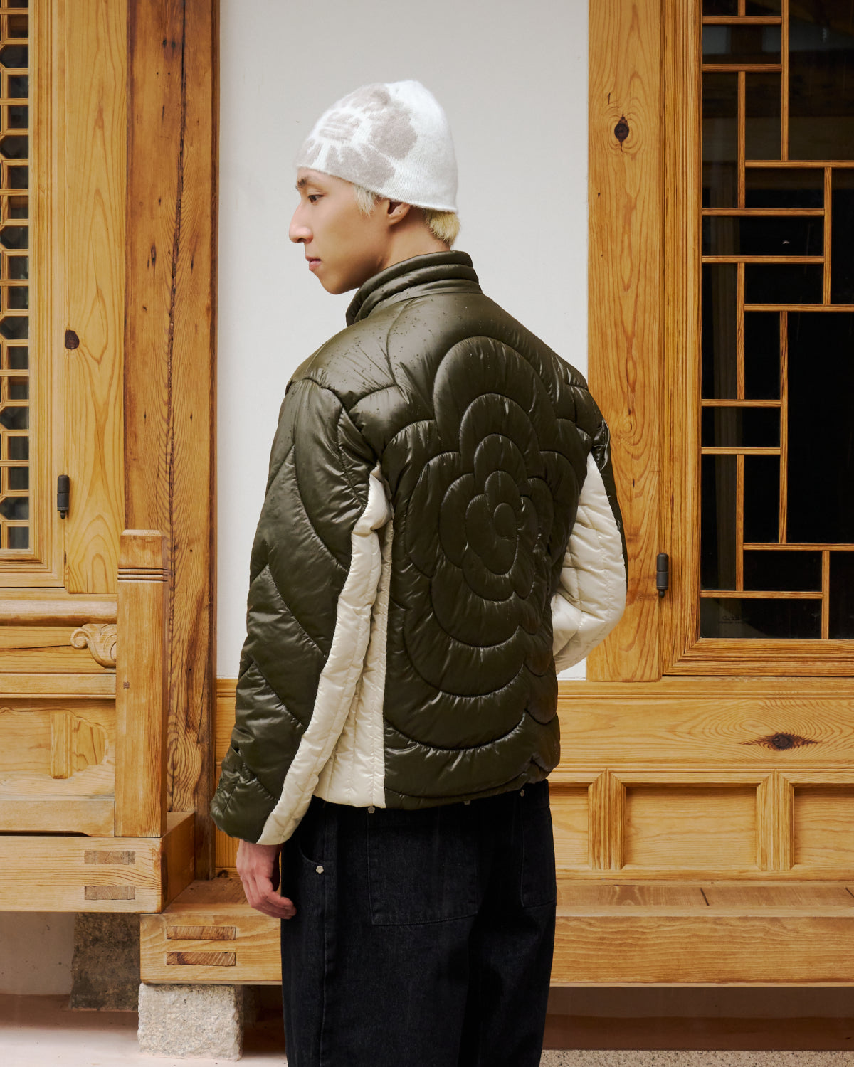 A person in a white knit beanie and the Sundae School OLIVE OATMEAL MOCHI SPIRAL PUFFER, featuring a bold spiral design on the back, stands with their back to a wooden building with detailed paneling and window.