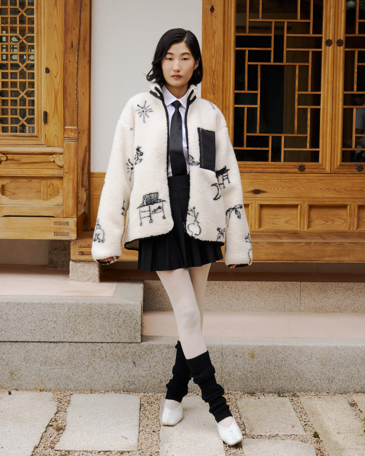 A young woman stands on stone steps in front of a traditional wooden building, wearing Sundae School’s Kiwa Fleece Zip-Up, paired with a black skirt, white tights, black leg warmers, white shoes, and a black tie over a white shirt.