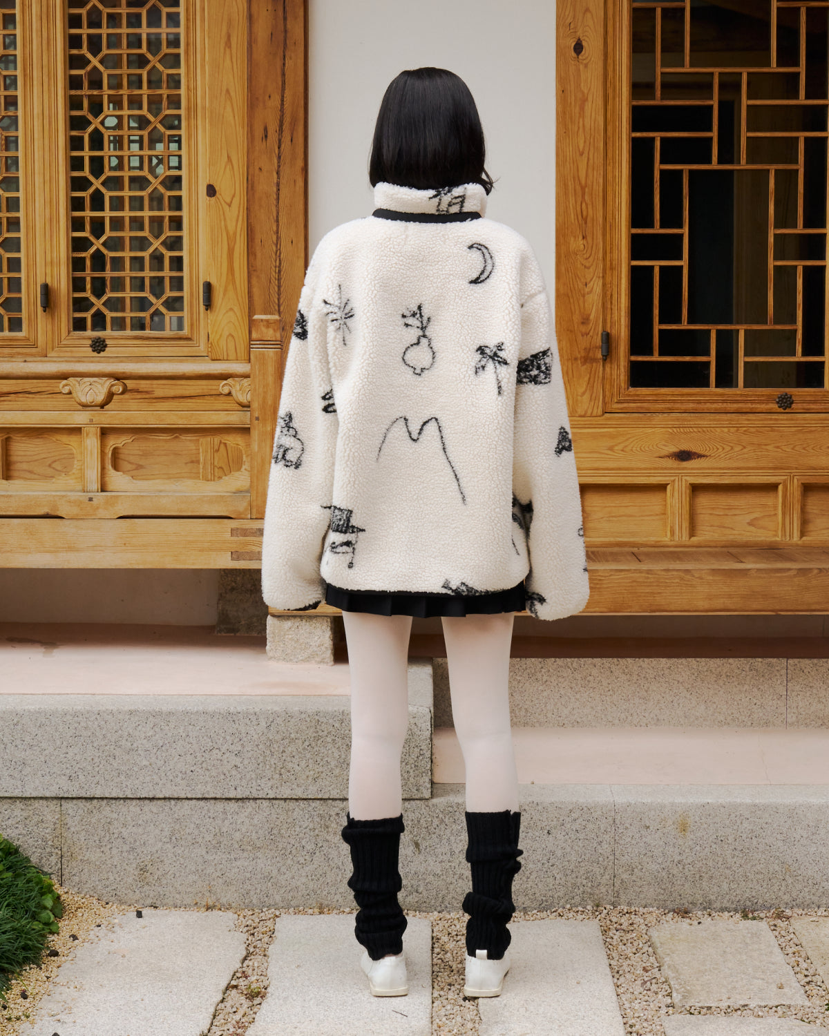 A person with short dark hair faces away, wearing a white Sundae School Kiwa Fleece Zip-Up with black abstract patterns, a short black skirt, white tights, black leg warmers, and white shoes in front of a traditional wooden building.