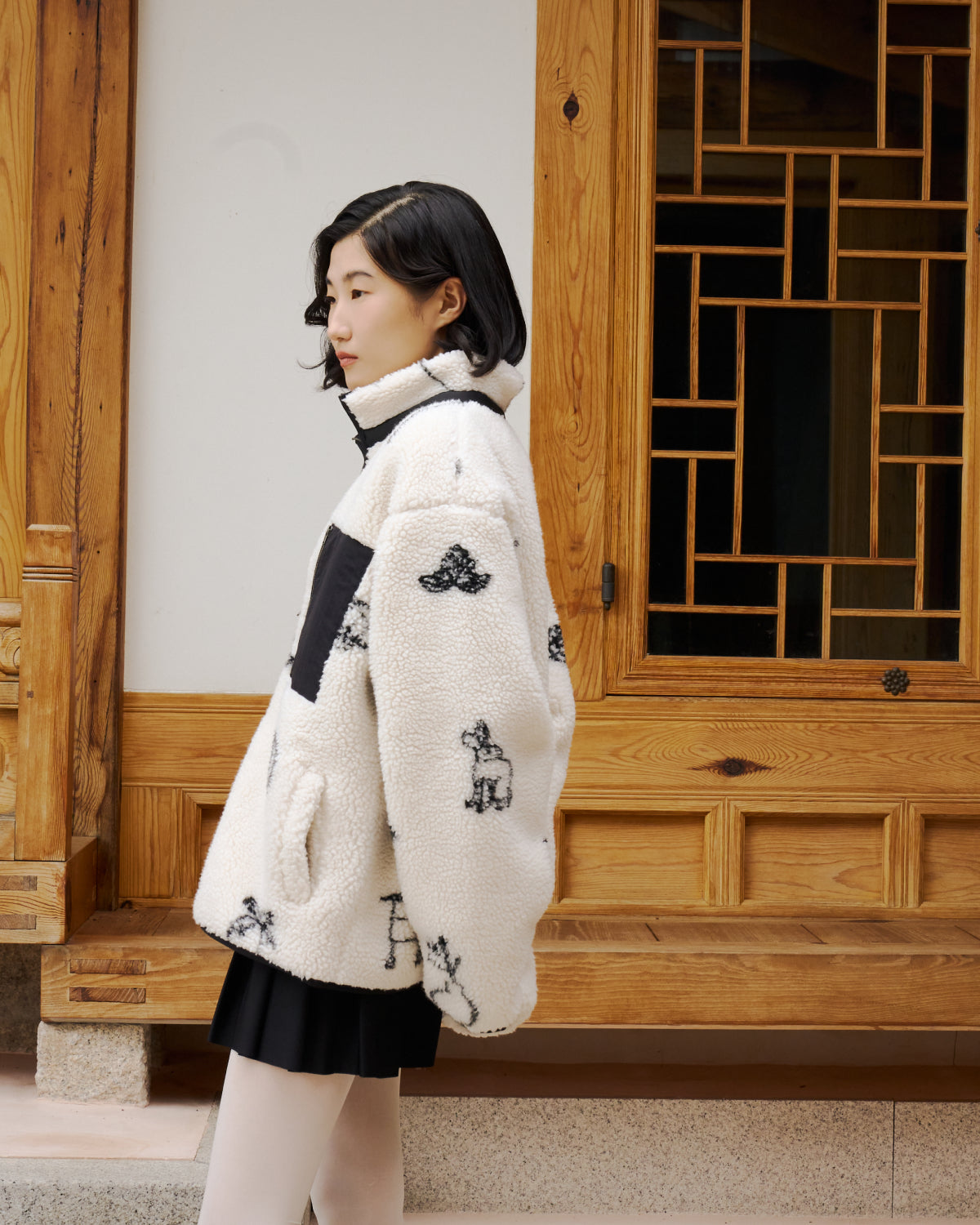 A woman with short dark hair stands sideways in front of a wooden door, wearing Sundae School's white Kiwa Fleece Zip-Up with black animal prints, a black skirt, and white tights.