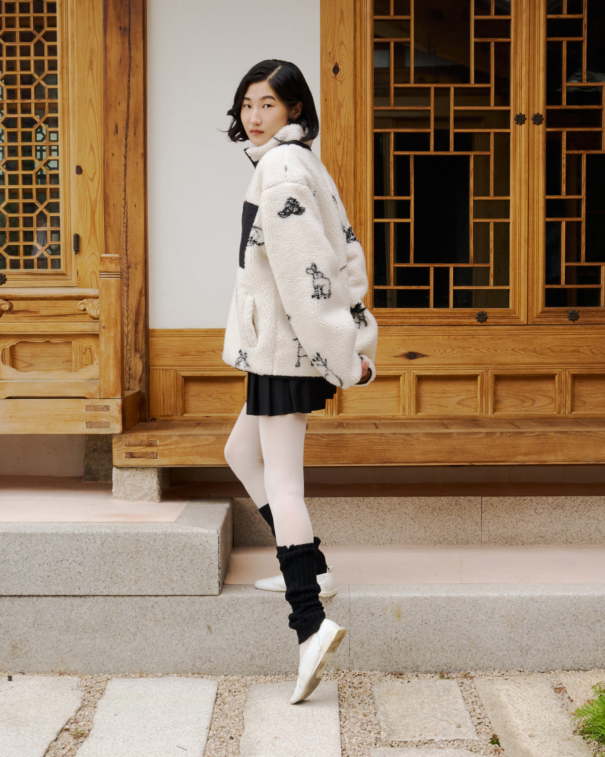 A woman with short dark hair stands on stone steps in front of a wooden building, wearing the Sundae School Kiwa Fleece Zip-Up with black designs, a black skirt, white tights, black leg warmers, and white shoes.