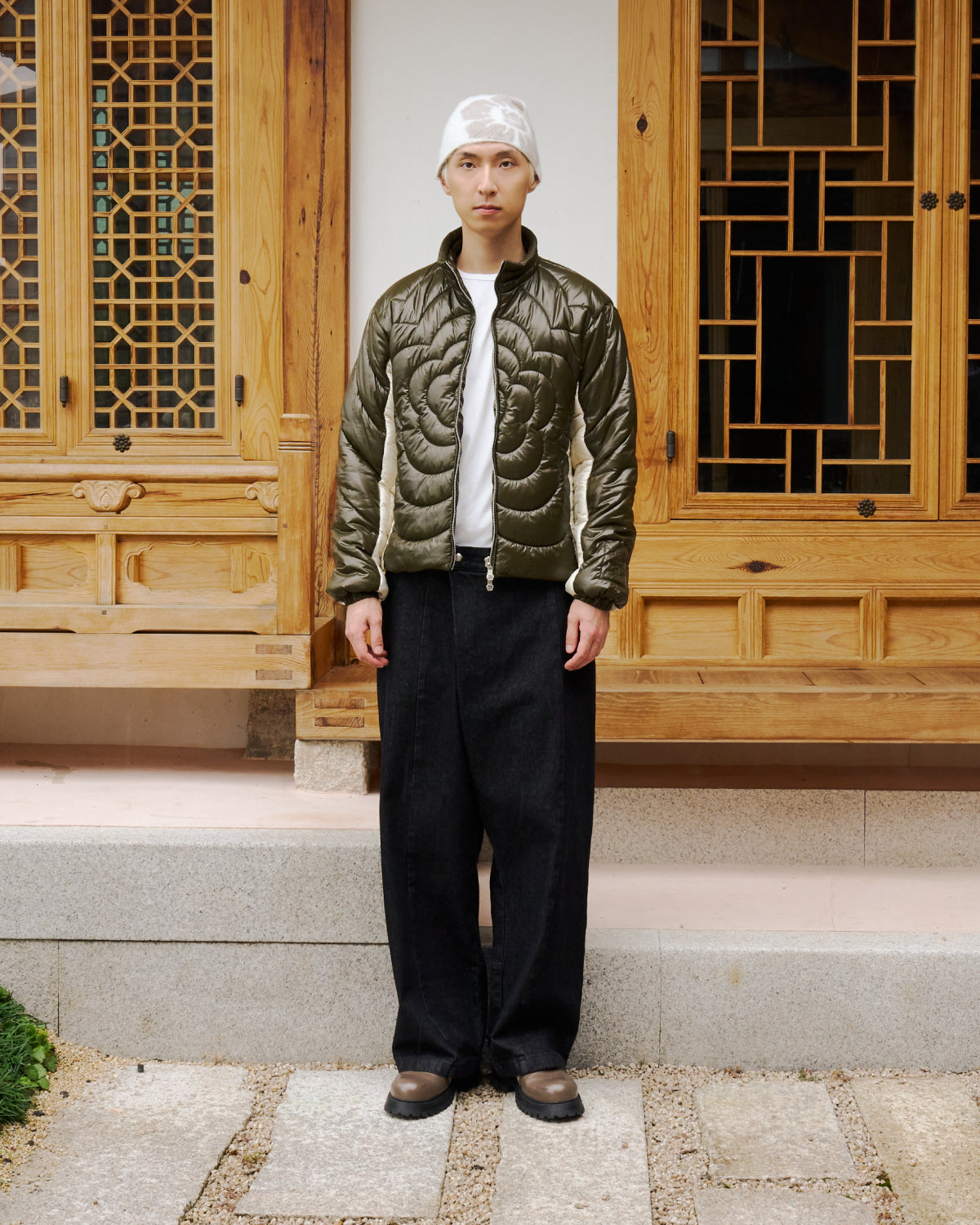 A person stands on a stone path in front of a wooden building, wearing the OLIVE OATMEAL MOCHI SPIRAL PUFFER by Sundae School, paired with a white shirt, wide black pants, brown shoes, and a white cap. Their expression is neutral with arms at their sides.