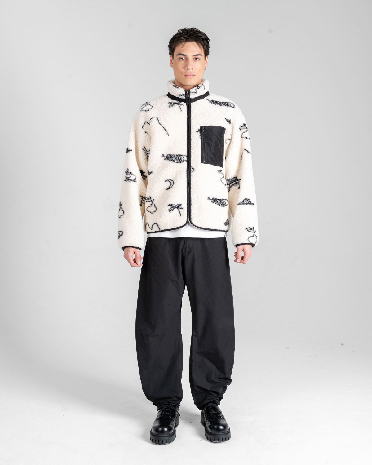 A man faces forward in a cream Sundae School Kiwa Fleece Zip-Up with black abstract patterns and a black chest pocket, paired with loose black pants and chunky black shoes, against a plain light gray background.
