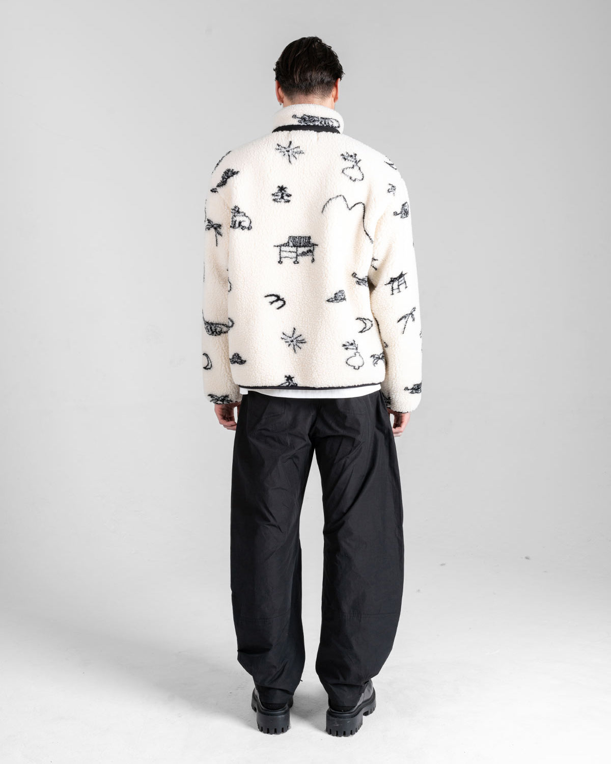 A person faces away, wearing the Sundae School Kiwa Fleece Zip-Up with black abstract patterns and black loose pants, in a minimal light gray studio.