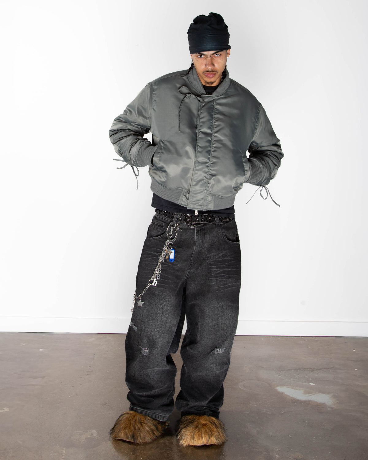 A person stands against a white wall, wearing the Sundae School KHAKI FLOWER CHANNELED BOMBER with adjustable flower sleeves, a black beanie, baggy black jeans with keychains, and fuzzy brown slippers. Their hands are tucked in the bomber’s pockets.