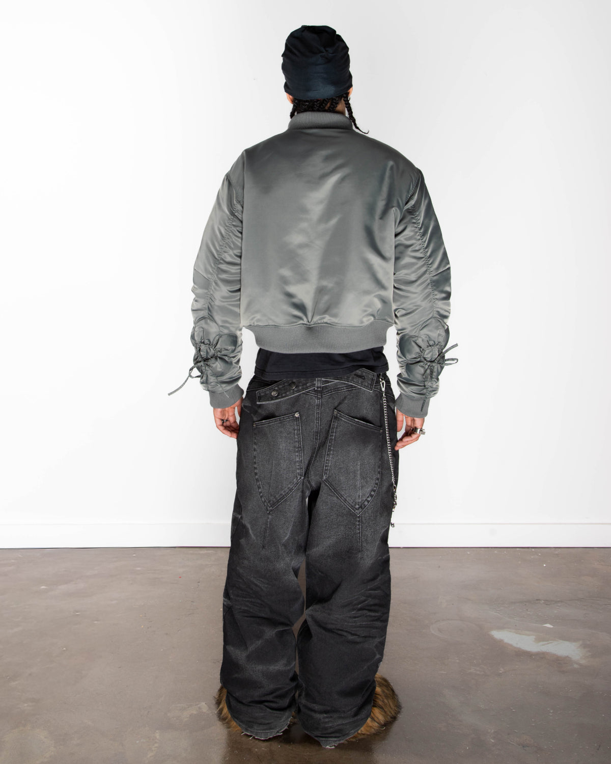 A person stands with their back to the camera, wearing Sundae School’s KHAKI FLOWER CHANNELED BOMBER with adjustable flower sleeves, loose black jeans, fur-trimmed shoes, and a dark head covering against a plain white wall and concrete floor.