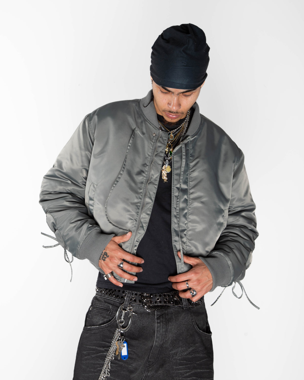 A person wears a khaki Sundae School Flower Channeled Bomber with adjustable flower sleeves, black beanie and shirt, jeans with a chain, rings, necklaces, visible tattoos, and looks down with hands on their waistband.