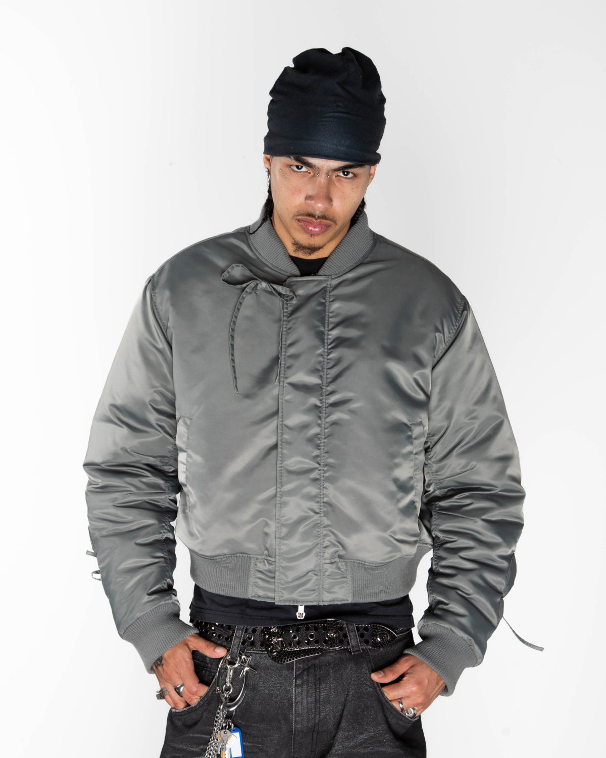 A person wears a Sundae School KHAKI FLOWER CHANNELED BOMBER with adjustable flower sleeves, a black beanie, and dark pants, standing against a white background with hands in pockets and a serious expression.