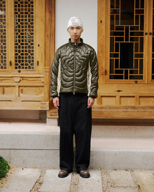 A person stands on stone steps before a wooden building with geometric windows, wearing Sundae School's OLIVE OATMEAL MOCHI SPIRAL PUFFER, black wide-leg pants, dark shoes, and a white cap.