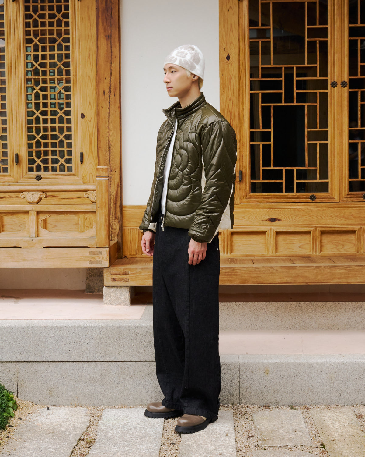 A man in the Sundae School OLIVE OATMEAL MOCHI SPIRAL PUFFER stands sideways before ornately carved wooden doors, styled with loose black pants, brown shoes, and a white patterned beanie.