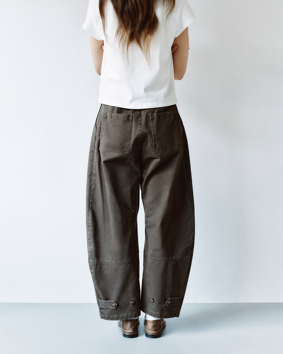 Back view of a person wearing olive green, high-waisted, wide-leg SOIL HANBOK CHINO PANTS with back patch pockets and adjustable button tabs at the cuffs.