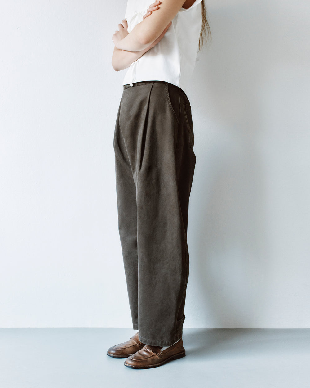 A person wearing a white top and SOIL HANBOK CHINO PANTS in dark gray with front pleats, standing in profile in brown loafers.