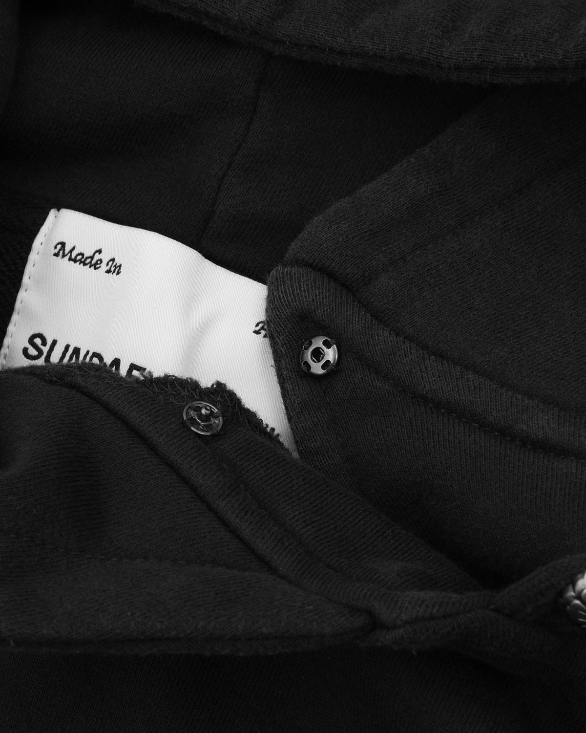 Close-up of the BLACK WASH HANBOK HOODIE by Sundae School, highlighting its oversized hood and a unique skull-shaped metal snap button. A partially visible white clothing label beneath the fabric displays printed text, including 