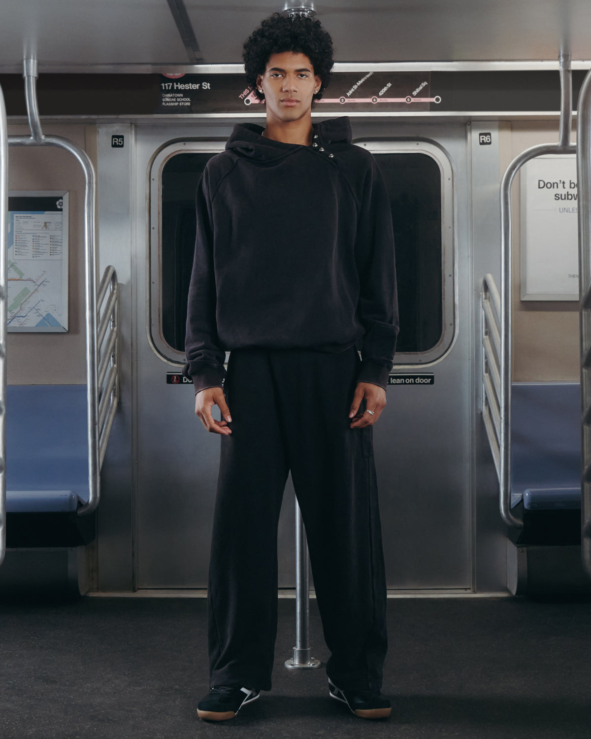 A person with curly hair stands in the middle of an empty subway car, facing the camera and wearing Sundae School's BLACK WASH HANBOK HOODIE—featuring an oversized hood—paired with black pants and black shoes.