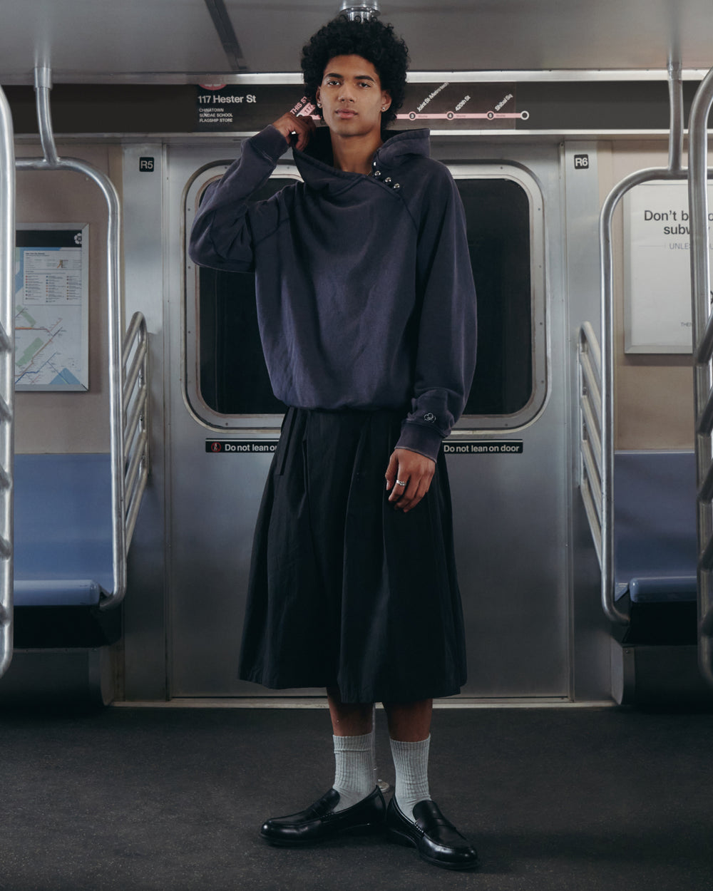 A person stands in a subway car wearing a dark oversized sweater, the Sundae School ASYMMETRIC CHIMA CULOTTE, white socks, and black shoes. The subway seats are empty and signs are visible on the walls behind them.