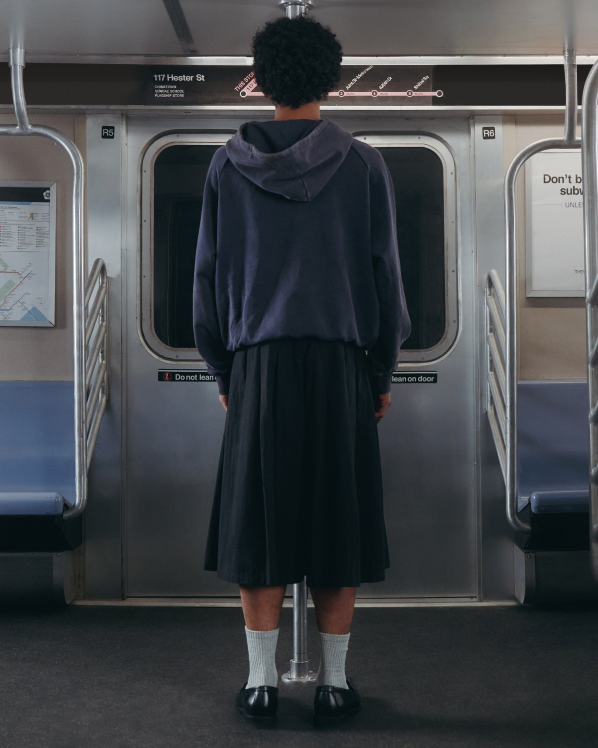 A person wearing a dark hoodie and Sundae School’s BLACK PLEATED HANBOK CULOTTES stands facing the closed doors on an empty subway train, with their back to the camera.