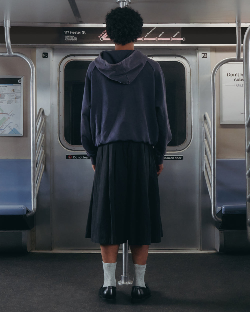 A person with short curly hair, dressed in the Sundae School NAVY WASH HANBOK HOODIE featuring an oversized hood, along with a long skirt, white socks, and black shoes, stands facing the subway doors in an empty subway car.